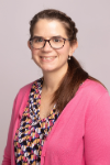 Woman with long brown hair and glasses wearing a pink sweater and floral shirt.