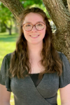 Young woman with long brown hair and glasses standing in front of a tree.
