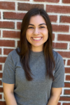 Young woman with longer dark brown hair standing in front of a brick wall.